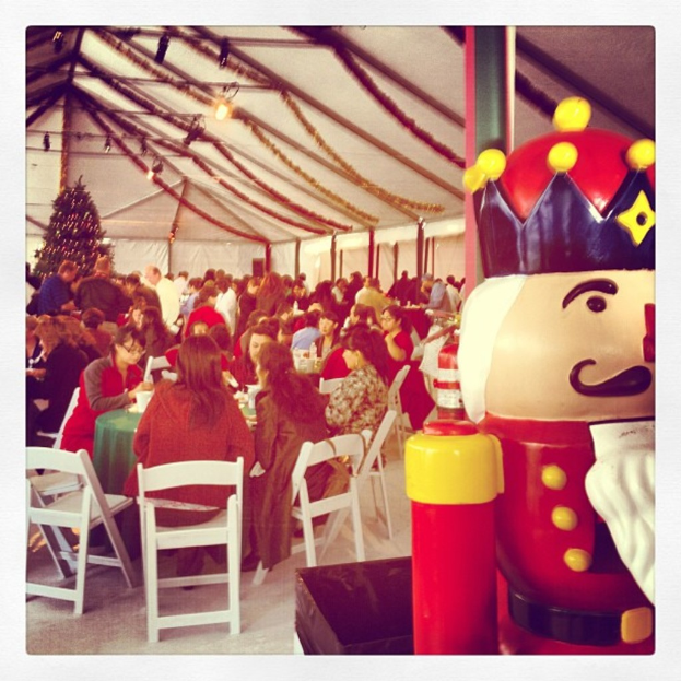 Eine Gruppe von Menschen feiert Weihnachten in einer Hütte, mit einem geschmückten Baum mit Lichtern auf dem Dach, weißen Stühlen, einem roten Spielzeug-Sicherheitswächter und einer schwarzen Krone.