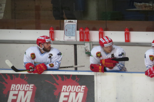 Drei Hockey-Spieler in Helmen und Handschuhen sitzen auf einer Bank und halten Hockey-Schläger, mit einem Text-Schild an der Wand dahinter und verschiedenen Gegenständen auf dem Podest, sowie einer Glaswand im Hintergrund.