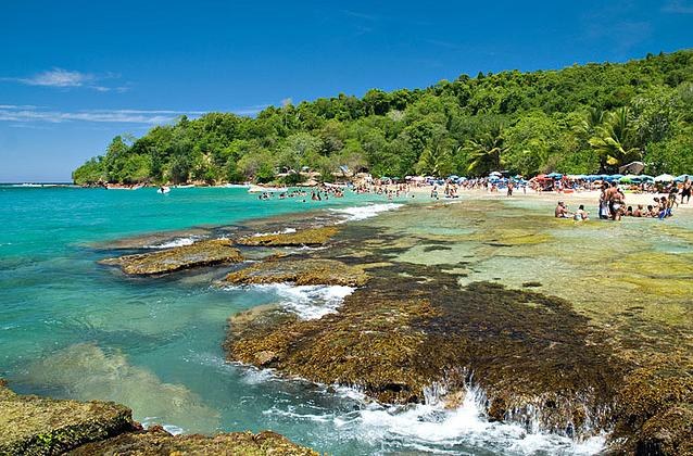 Ein überfüllter Strand mit Menschen auf einem felsigen Ufer, beschattet von Sonnenschirmen, vor Bäumen und einem bewölkten Himmel in Phuket, Thailand.
