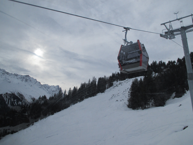 Eine Seilbahn fährt einen schneebedeckten Berg hinauf mit Bäumen im Hintergrund, unter einem bewölkten Himmel mit hellerer Sonne.