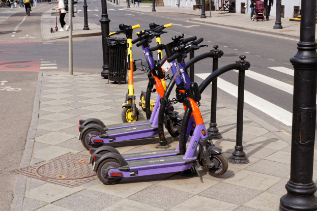 Eine Reihe von Elektro-Scootern, die an einer Stra├če geparkt sind, mit Fü├čgängern, Radfahrern, einem Müllcontainer im Vordergrund und Geb├Ąuden, B├Ąumen und Pflanzen im Hintergrund.