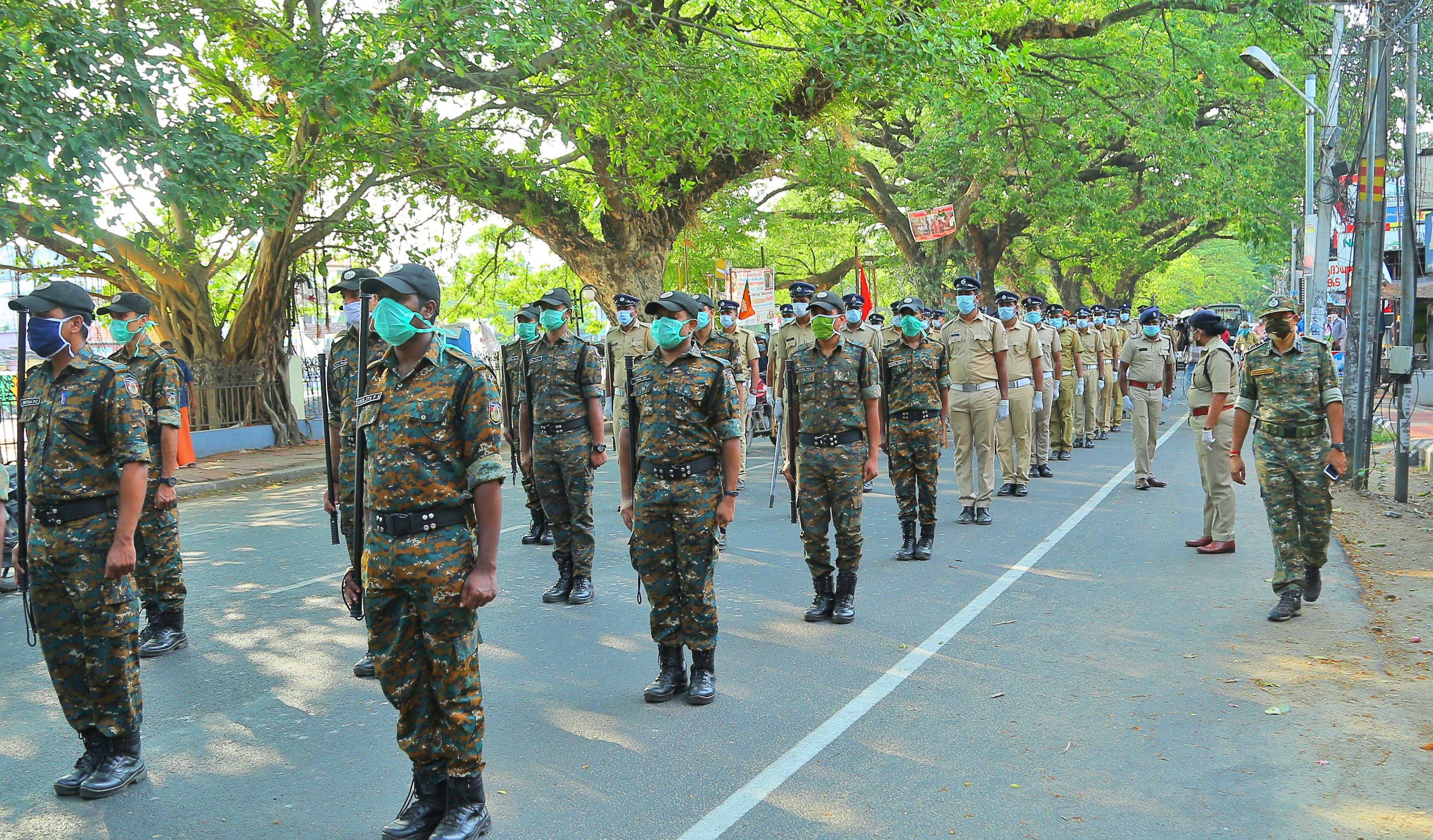 A group of police officers in uniform, caps, and masks walking down a tree-lined street with light poles and text boards, with buildings and a clear blue sky in the background.