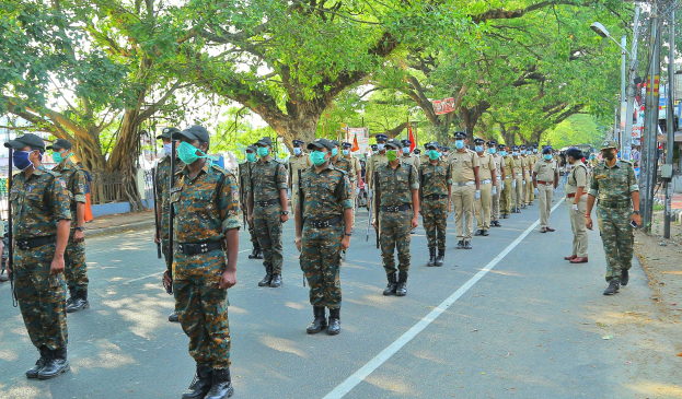 A group of police officers in uniform, caps, and masks walking down a tree-lined street with light poles and text boards, with buildings and a clear blue sky in the background.