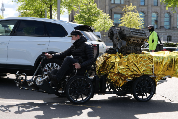Ein Mann im Rollstuhl mit einem großen Motor daran befestigt, umgeben von Fahrzeugen auf einer Straße mit Bäumen, Gebäuden und einem klaren blauen Himmel im Hintergrund; er trägt eine schwarze Jacke, eine Kappe und hält ein Objekt in der Hand.