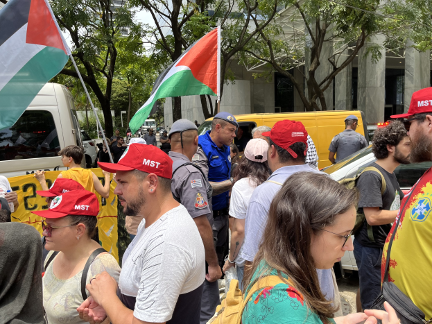 Eine Gruppe von Menschen steht vor einem Gebäude während einer Demonstration, einige tragen Mützen und Taschen, mit Fahrzeugen auf der Straße und Bäumen im Hintergrund bei einem klaren blauen Himmel, mehrere halten palästinensische Flaggen.