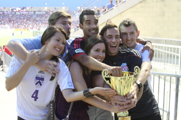 Sechs Menschen, darunter zwei Frauen und vier Männer, halten einen Pokal und lächeln, während sie für ein Foto vor einem Eisengeländer posieren, mit einer Wand und einem Geländer im rechten oberen Eck, Menschen und Lichter im Hintergrund, möglicherweise in einem Stadion.