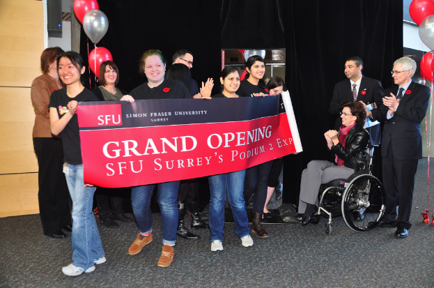 Eine Gruppe Frauen hält ein Banner hoch, um ein Ereignis zu bewerben, mit zwei klatschenden Männern daneben und einer sitzenden, aufgeregten Frau im Rollstuhl vor einem schwarzen Vorhang und Ballons.