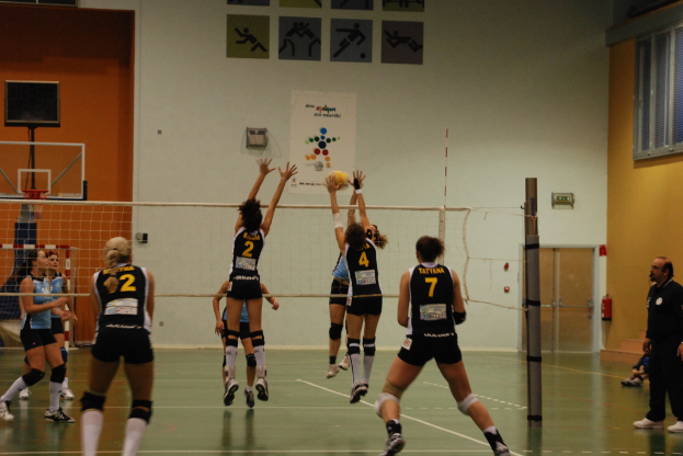 Ein Volleyballspiel auf einem Indoor-Court mit Spielern, einem Basketball-Netz und einer Tür und Fenstern im Hintergrund.