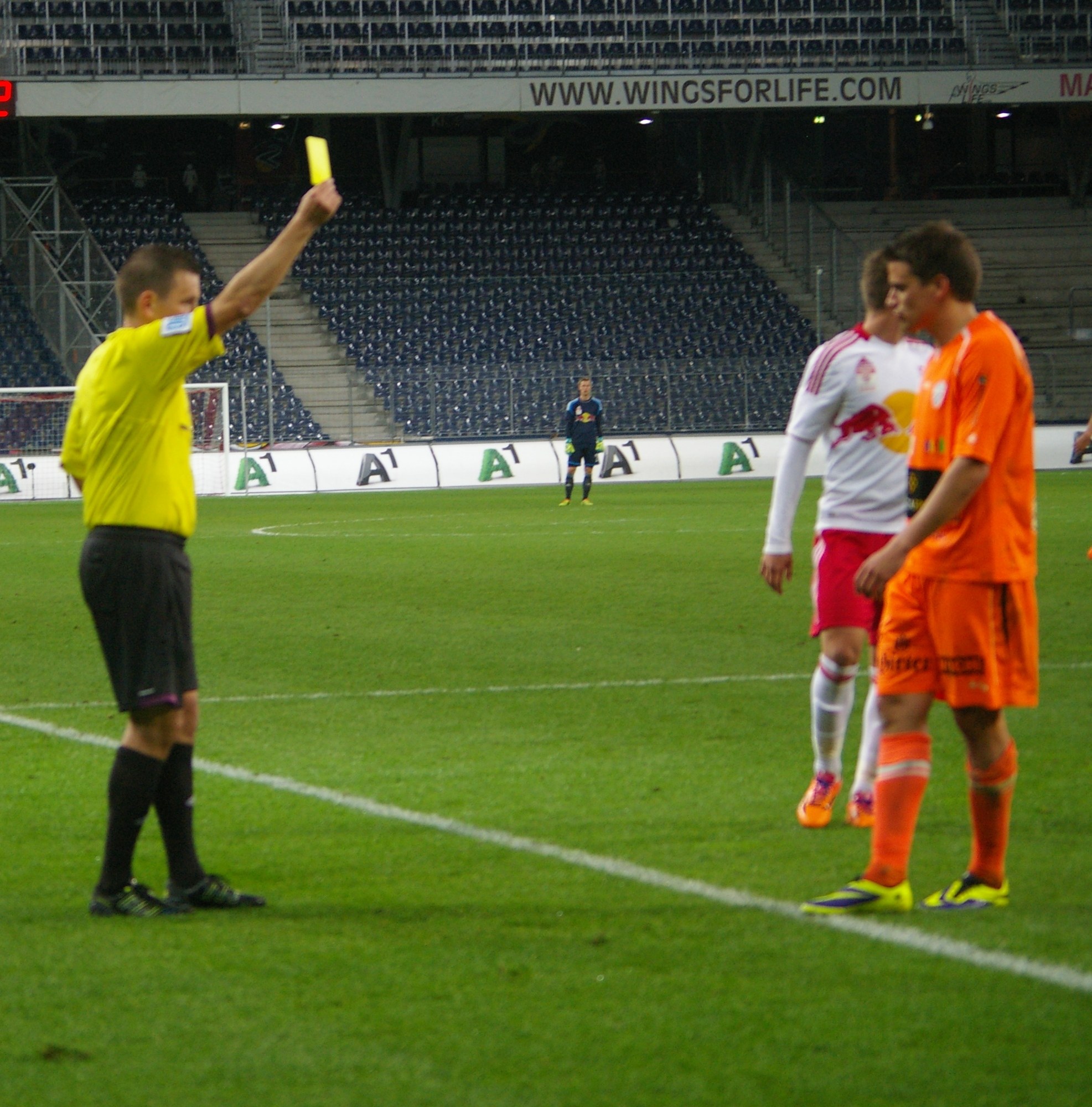 A referee issuing a yellow card to a soccer player on the field, with a goal post, fence, signboard, metal frames, display screen, chairs, and ceiling lights visible in the background.