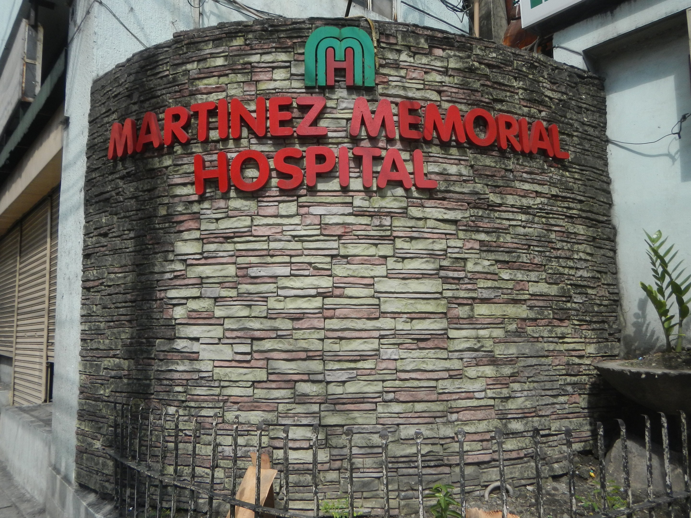 Ein mehrstöckiges Krankenhausgebäude namens "Martinez Memorial Hospital" in Manila, Philippinen, umgeben von einem Metallzaun, Blumentöpfen und einem bewölkten Himmel.