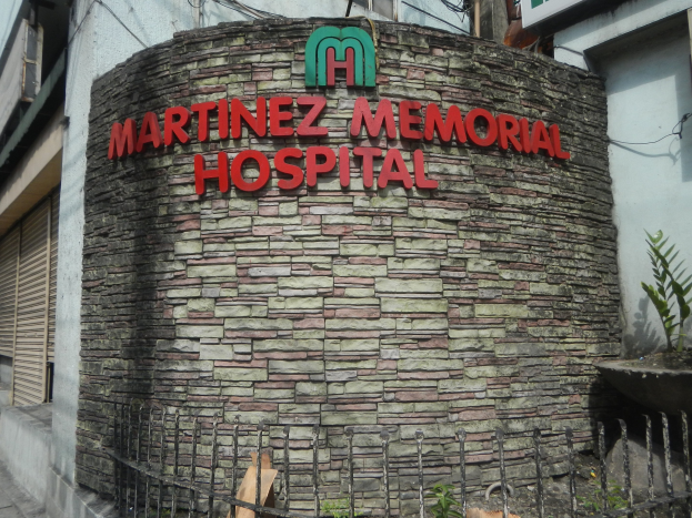 Ein mehrstöckiges Krankenhausgebäude namens "Martinez Memorial Hospital" in Manila, Philippinen, umgeben von einem Metallzaun, Blumentöpfen und einem bewölkten Himmel.