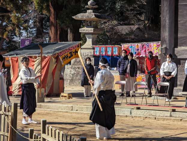 Eine Gruppe von Menschen in traditioneller Kleidung, einige mit Masken und Stöcken, versammelt sich vor einem Gebäude während einer Zeremonie in Kyoto, mit Stühlen, Bannern, einem Zelt und Bäumen unter einem klaren blauen Himmel.