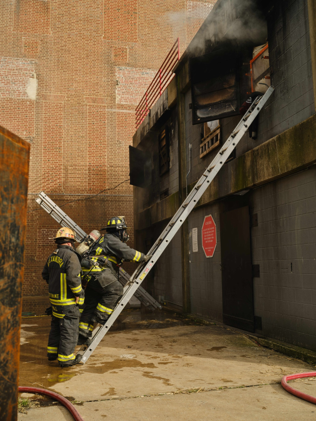 Feuerwehrleute in Helmen klettern eine Leiter hinauf zu einem raucherfüllten Gebäude, mit einem Metallobjekt links und Rohren am Boden.