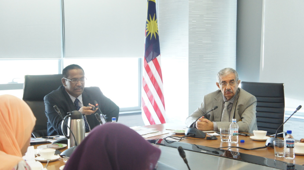 Eine Gruppe von Menschen, darunter Männer in Anzügen und zwei Frauen in Burkas, sitzt um einen Tisch mit Wasserflaschen, einem Mikrofon, Tassen und einem Teekessel, mit einem Fenster und einer Flagge im Hintergrund.