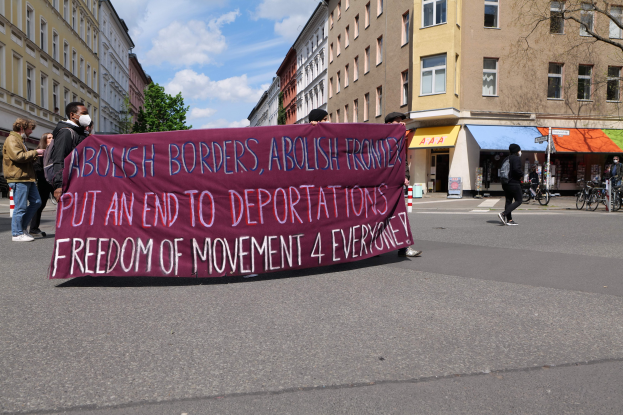 Eine Gruppe von Menschen marschiert auf einer Stadtstraße mit einem Banner, auf dem "Abolish Borders, Abolish Frontiers, Put an End to Deportations, Freedom of Movement 4 Everyone" steht, mit Gebäuden, Bäumen, Fahrrädern und bewölktem Himmel im Hintergrund.