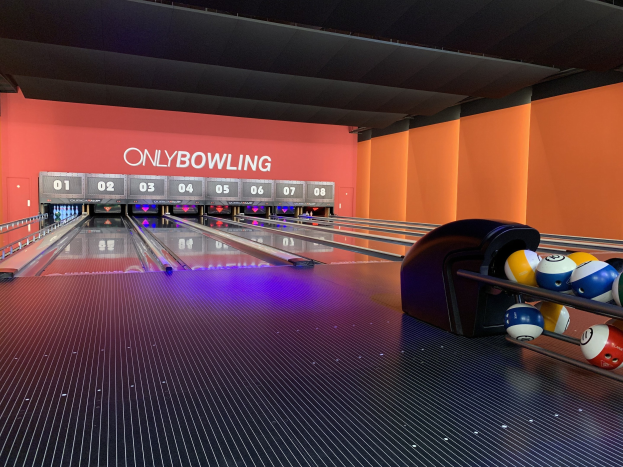 Eine Bowlingbahn mit einer Reihe von Bowlingkugeln auf der rechten Seite, aufgestellte Bowlingpins in der Mitte und eine Wand mit Text im Hintergrund, mit der Decke oben sichtbar.