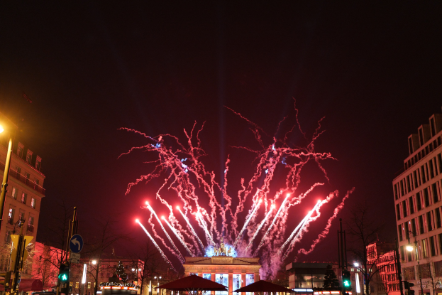 Eine belebte Stadtstraße in Berlin an Silvester, voller Menschen, Fahrzeuge und Gebäude, beleuchtet von Lichtern und Feuerwerk am Himmel.