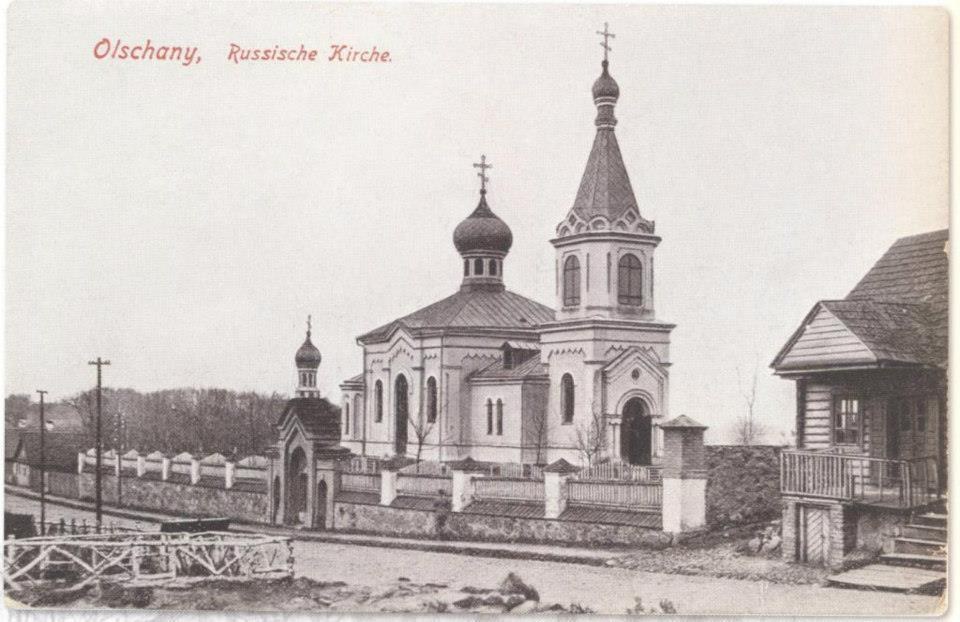 Schwarz-weiß-Fotografie einer russisch-orthodoxen Kirche in Olschany, Russland, umgeben von Häusern, Bäumen, Strommasten und einem Zaun unter einem klaren Himmel, mit Text oben.