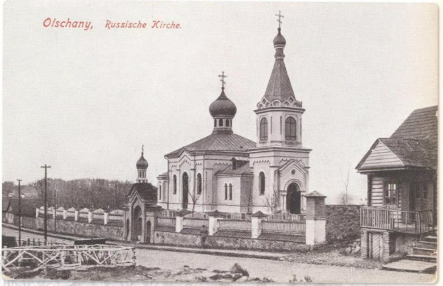 Schwarz-weiß-Fotografie einer russisch-orthodoxen Kirche in Olschany, Russland, umgeben von Häusern, Bäumen, Strommasten und einem Zaun unter einem klaren Himmel, mit Text oben.