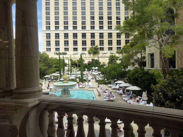 Eine Gruppe von Menschen, die auf Bänken auf einem Balkon mit weißen Säulen und einem Balustraden-Geländer sitzen, mit Bäumen, Pflanzen und einem großen Gebäude mit Glasfenstern im Hintergrund.