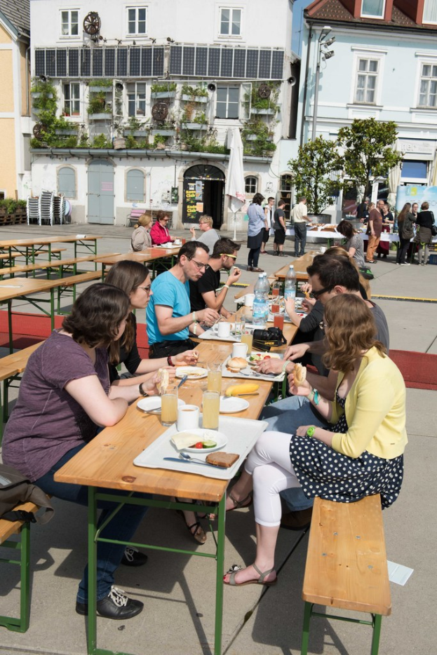 Menschen sitzen auf Bänken vor einem Tisch mit Löffeln, Gläsern, Essen und Wasserflaschen, mit Gebäuden und Pflanzen im Hintergrund und einem Schirm in der Mitte.