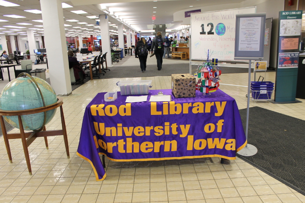 Eine Ausstellung mit einem Tisch, auf dem verschiedene Landesflaggen, eine Box, Papiere und ein Banner liegen, eine Kugel zur Linken, eine Anschlagtafel zur Rechten mit einigen Menschen in der Nähe und mehrere Tische und Stühle ganz links.