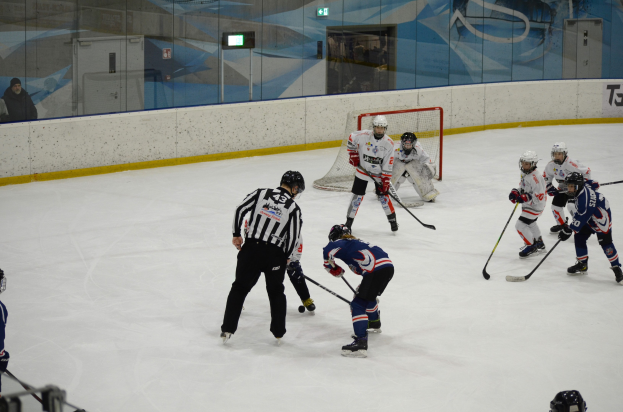Gruppe von Menschen, die Eis-Hockey auf einem Eisplatz spielen, mit Helmen und Hockey-Schlägern, einem Netz in der Mitte, Zuschauern auf der linken Seite und einer Werbung an der Wand im Hintergrund.