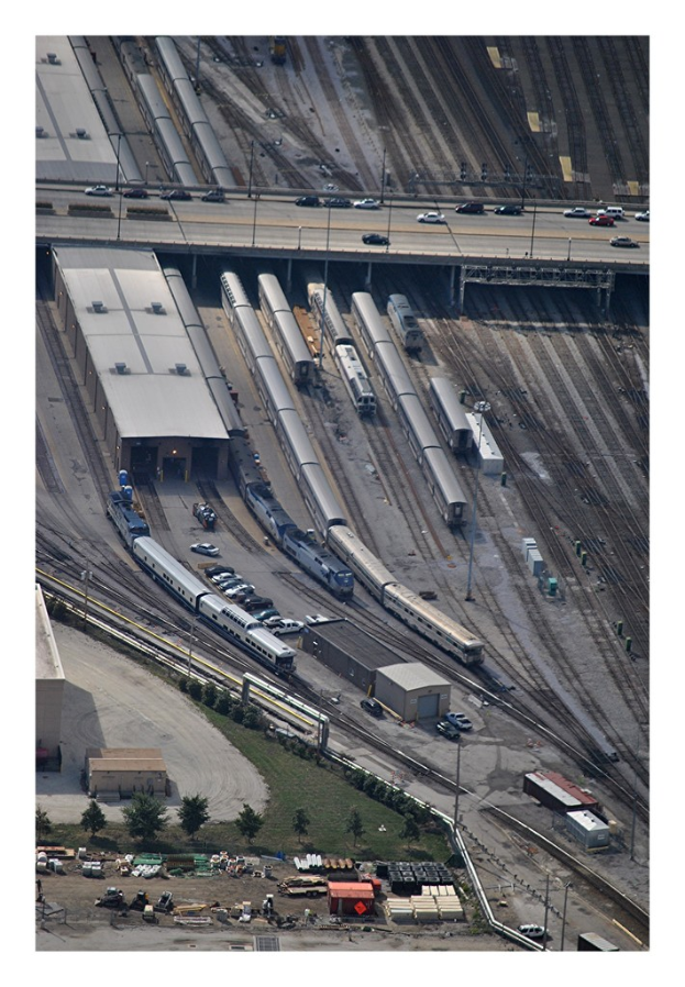 Eine Brücke mit wenigen Fahrzeugen darauf, Eisenbahnschienen mit wenigen Zügen darunter und eine Landschaft mit Gebäuden, Bäumen, Pfählen und verschiedenen Gegenständen und Fahrzeugen am Boden.