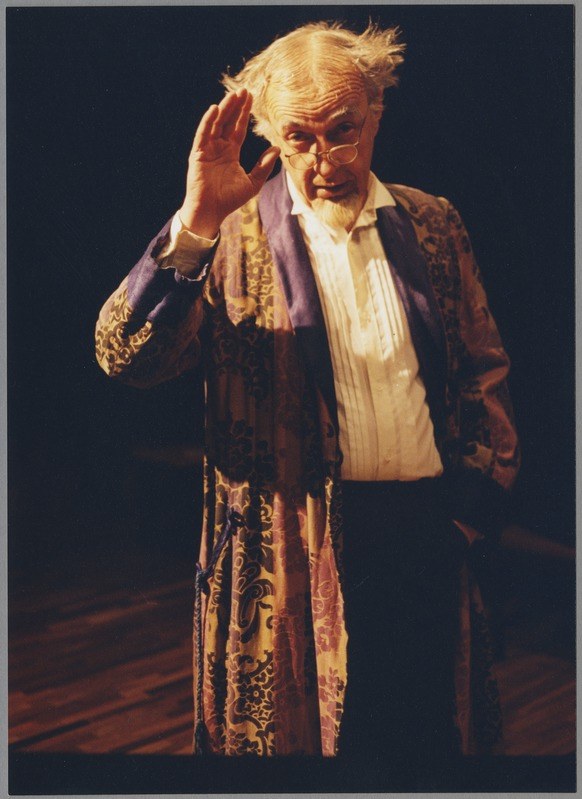 A man in a long robe and glasses stands on a stage with clasped hands, facing an audience against a dark background.