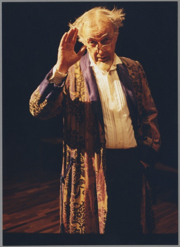 A man in a long robe and glasses stands on a stage with clasped hands, facing an audience against a dark background.