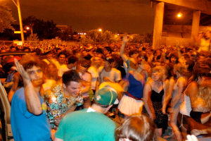 Menschen tanzen im Vordergrund eines Konzerts mit Lichtern, Stangen, Bäumen und anderen Objekten im Hintergrund.