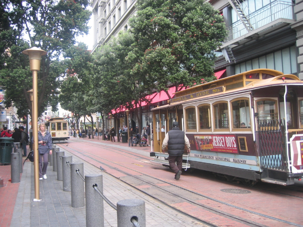 Eisenbahnschienen auf einer Straße mit Zügen, eine Person in der Nähe, kleine Pfosten mit Seilen auf der linken Seite, Straßenlaternen, Bäume, viele Menschen, ein Müllcontainer auf der linken Seite und Gebäude mit Leitern auf der rechten Seite.