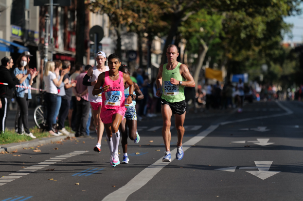 Gruppe von Läufern bei einem Marathon auf einer Stadtstraße mit Zuschauern auf der linken Seite, unscharfe Gebäude, Bäume und ein Fahrrad im Hintergrund.