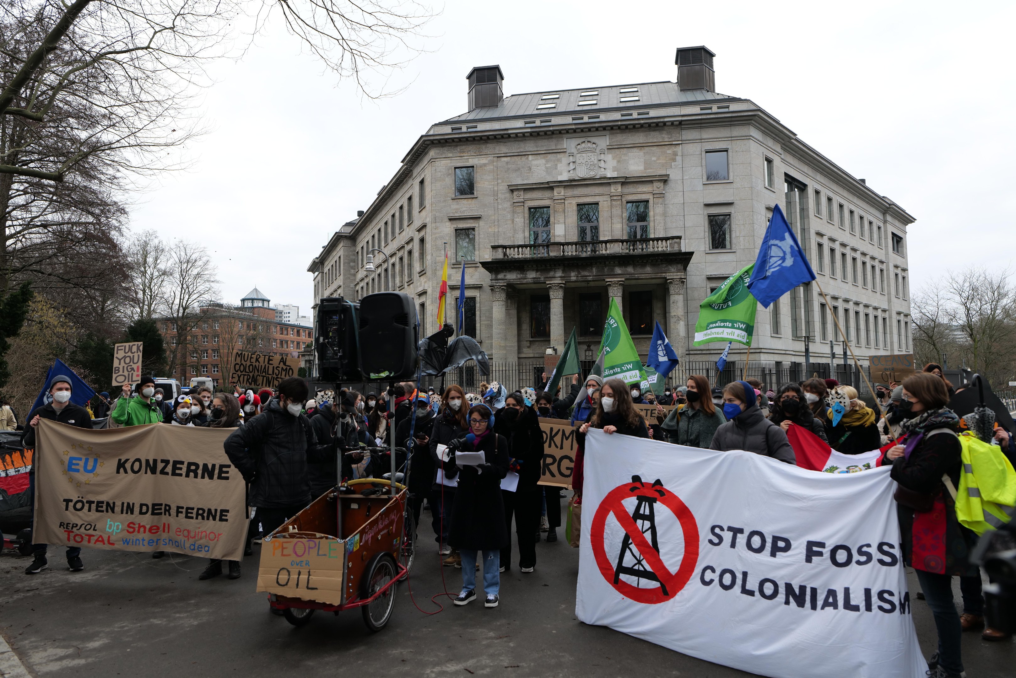 Large group of people marching in a protest against fossil fuels, carrying banners and flags, with a vehicle in the foreground, trees and buildings in the background under a clear blue sky.