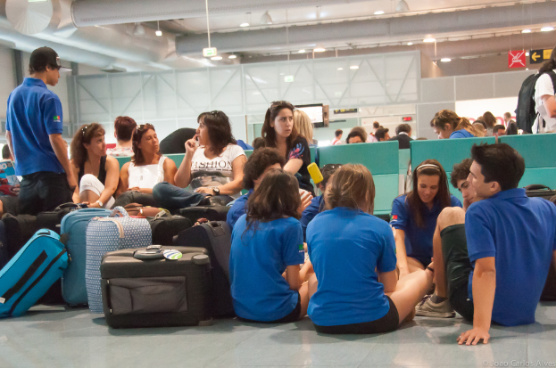 Ein belebtes Flughafeninnere mit vielen auf Flüge wartenden Menschen, Gepäck auf der linken Seite und Glastüren mit Richtungsschildern im Hintergrund.