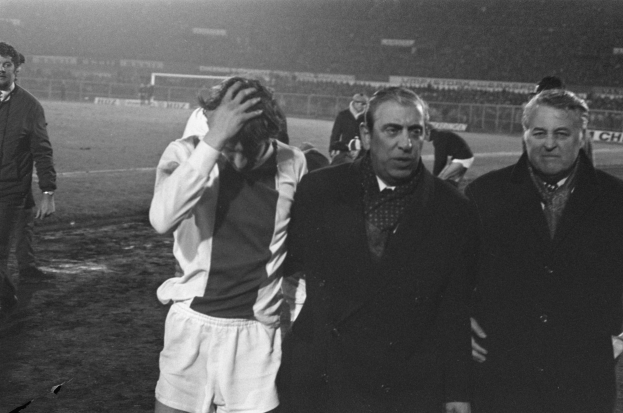 Schwarzes und weißes Bild einer Gruppe von Männern auf einem Fußballfeld, die nach einem Sieg enttäuscht wirken, mit einem Stadion im Hintergrund.