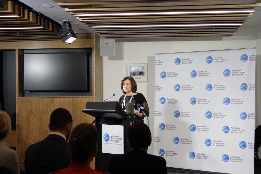 Eine Frau steht auf einer Bühne und spricht in ein Mikrofon, vor ihr sitzen Menschen auf Stühlen, an der Wand hängt ein Banner, an der hölzernen Wand steht ein Fernseher und an der Decke leuchten Lampen.