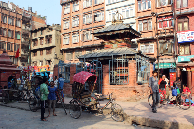 A bustling street in Kathmandu, Nepal, with people walking alongside tall buildings, rickshaws, bicycles, and other vehicles on the road, and people sitting and standing around under a clear blue sky.