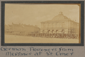 Eine Gruppe deutscher Kriegsgefangener aus Messines steht vor einem Gebäude an einem klaren blauen Himmel, mit Text am unteren Bildrand.