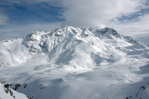 Ein schneebedeckter Berg mit ein paar Skifahrern, die darauf hinabfahren, umgeben von einer wolkenverhangenen Himmel und unberührtem Schnee.