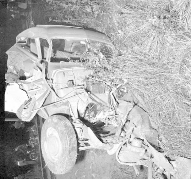Schwarz-weiß-Foto eines schwer beschädigten Autos mit eingedrückter Front und zersplitterten Fenstern am Straßenrand, mit einem Baum und verstreuten Gegenständen im Hintergrund.