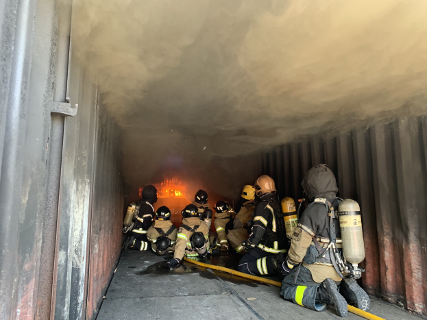 Feuerwehrleute mit Helmen und Sauerstoffflaschen arbeiten in einem raucherfüllten Tunnel, einer hält ein Rohr, mit sichtbarem Feuer und einer Wand auf der linken Seite.