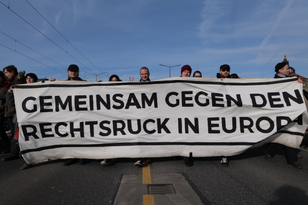 Eine Gruppe von Menschen steht auf der Straße und hält ein Schild mit der Aufschrift "Gemeinsam Gegen den Rechtsruck in Europa" hoch, im Hintergrund sind Straßenlaternen, Strommasten, Stromkabel, Gebäude und ein bewölkter Himmel zu sehen.