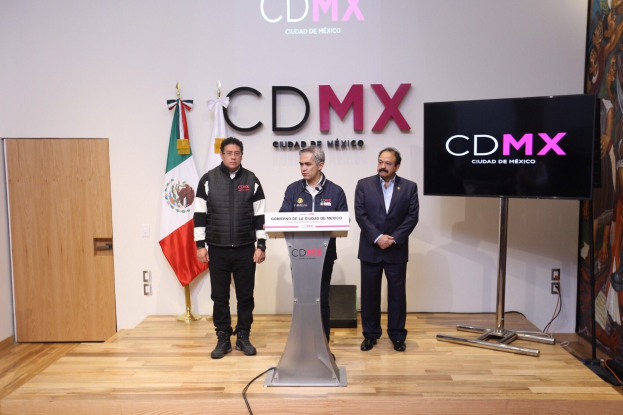 Drei Männer stehen auf einem Podium mit einem Mikrofon während einer Pressekonferenz in Mexiko, mit Flaggen, einer Tür und einem Banner im Hintergrund.