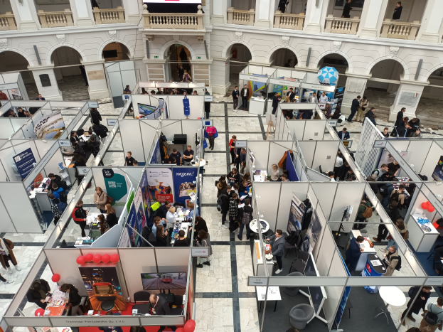 Große Besuchermenge auf einer internationalen Fachmesse mit Ständen, Bannern, Tischen, Stühlen und Luftballons in einem Saal mit gewölbter Architektur und einem großen Bildschirm im Hintergrund.