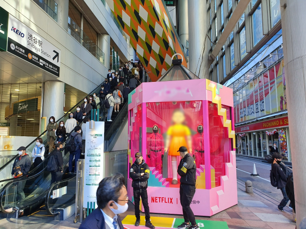 Eine Gruppe von Menschen steht vor einem Gebäude mit Glaswänden und Texttafeln, einige tragen Masken, in der Nähe einer Rolltreppe; ein pinker Kasten in der Mitte zeigt Schaufensterpuppen und trägt "Netflix's new Netflix booth in Tokyo."