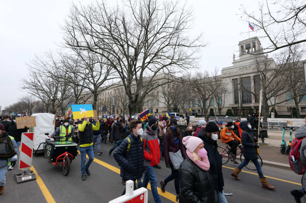 A large protest march in Washington, D.C. on January 21, 2020, with people walking, holding signs, and riding bicycles down a street in front of a building, under a clear blue sky.