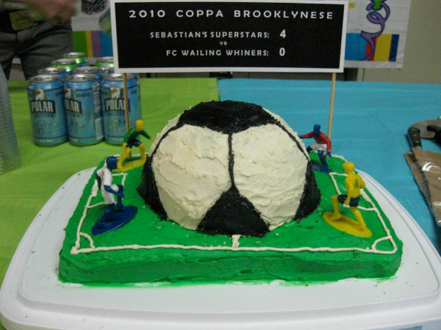 Ein fußballstadionförmiger Kuchen mit einem Fußball und vier Spielern, einer Fahne, Softgetränkedosen und einer Person links daneben, mit einer Tafel auf dem Kuchen.
