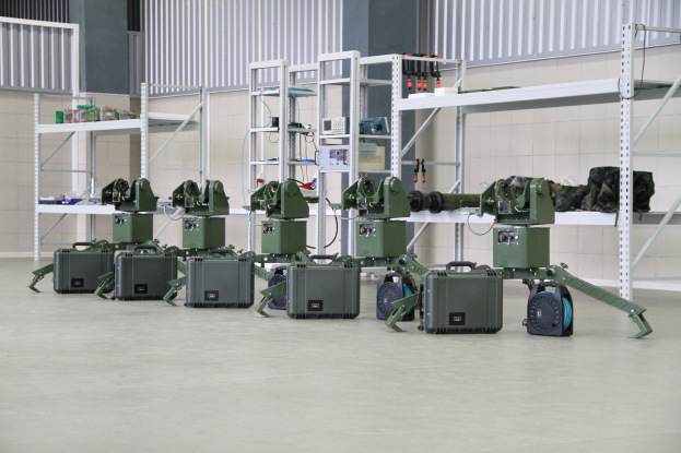 Eine Gruppe von Militärrobotern auf einem Boden mit Koffern und anderen Gegenständen auf umliegenden Gestellen, vor einer Wand mit einem Geländer im Hintergrund.