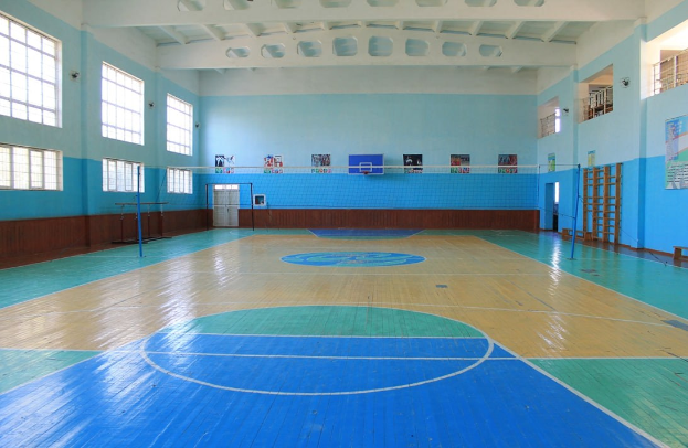 Große Turnhalle mit blau-weißem Boden, Basketballfeld, Netz, Stangen, einer Tür, Fenstern, Wandplakaten, Geländer und Dach.